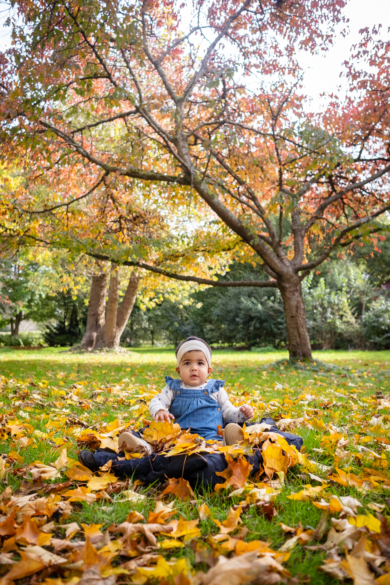 A baby sits among scattered autumn leaves in a park, wearing a denim romper and headband, with a background of vibrant orange and yellow foliage. The image captures a serene autumn moment, ideal for family photography ideas that highlight the colors and atmosphere of the fall season.