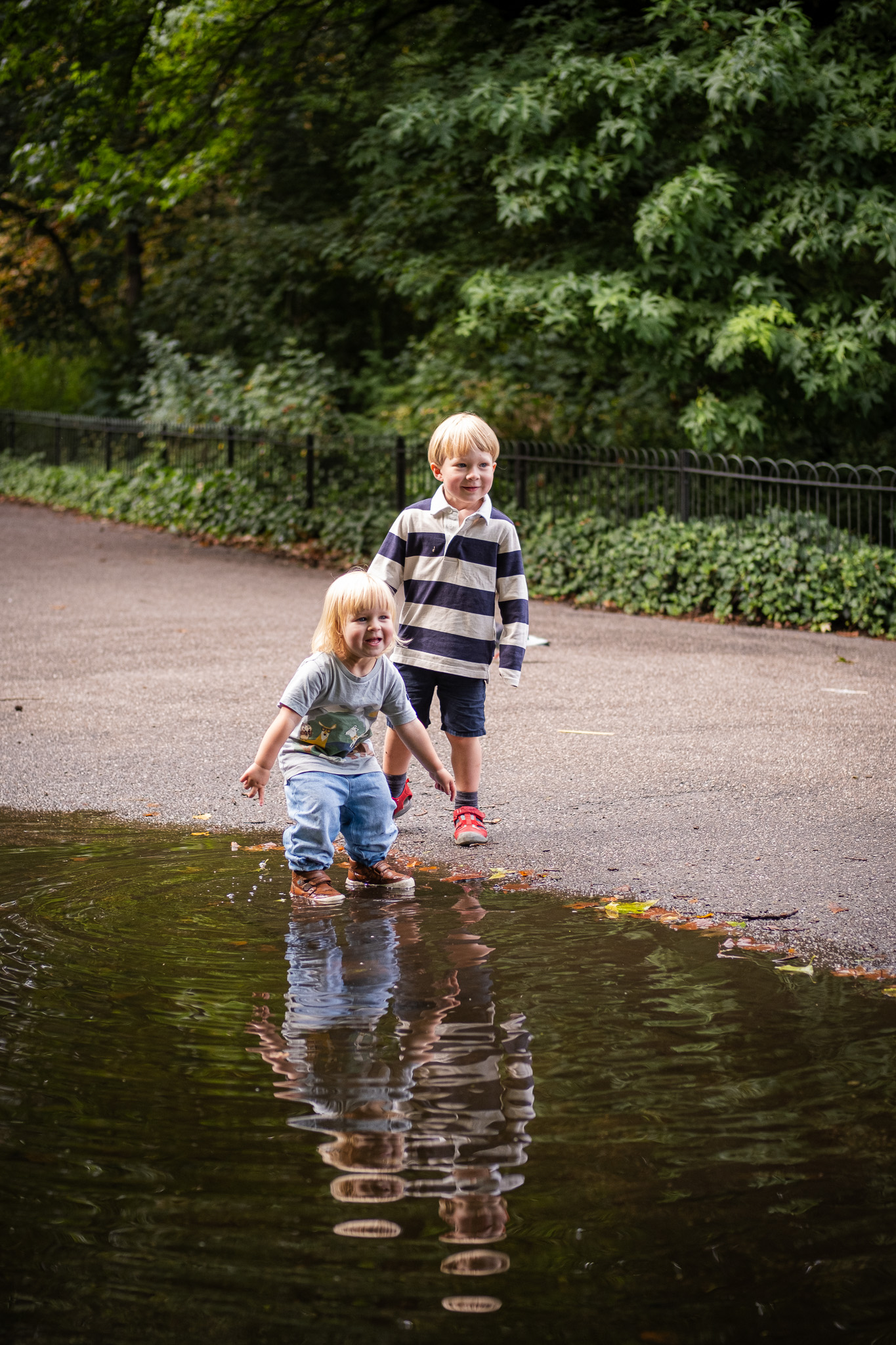 Two children play by the edge of a large puddle on a park path, with trees and greenery in the background.