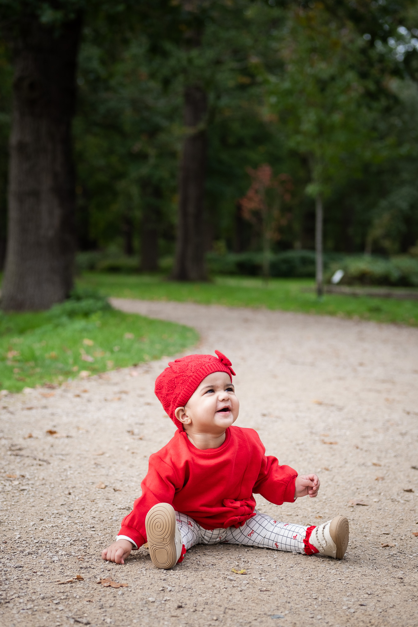 Little baby girl wearing red top and hat, sitting in the middle of a walking path, during autumn time at Bushy park