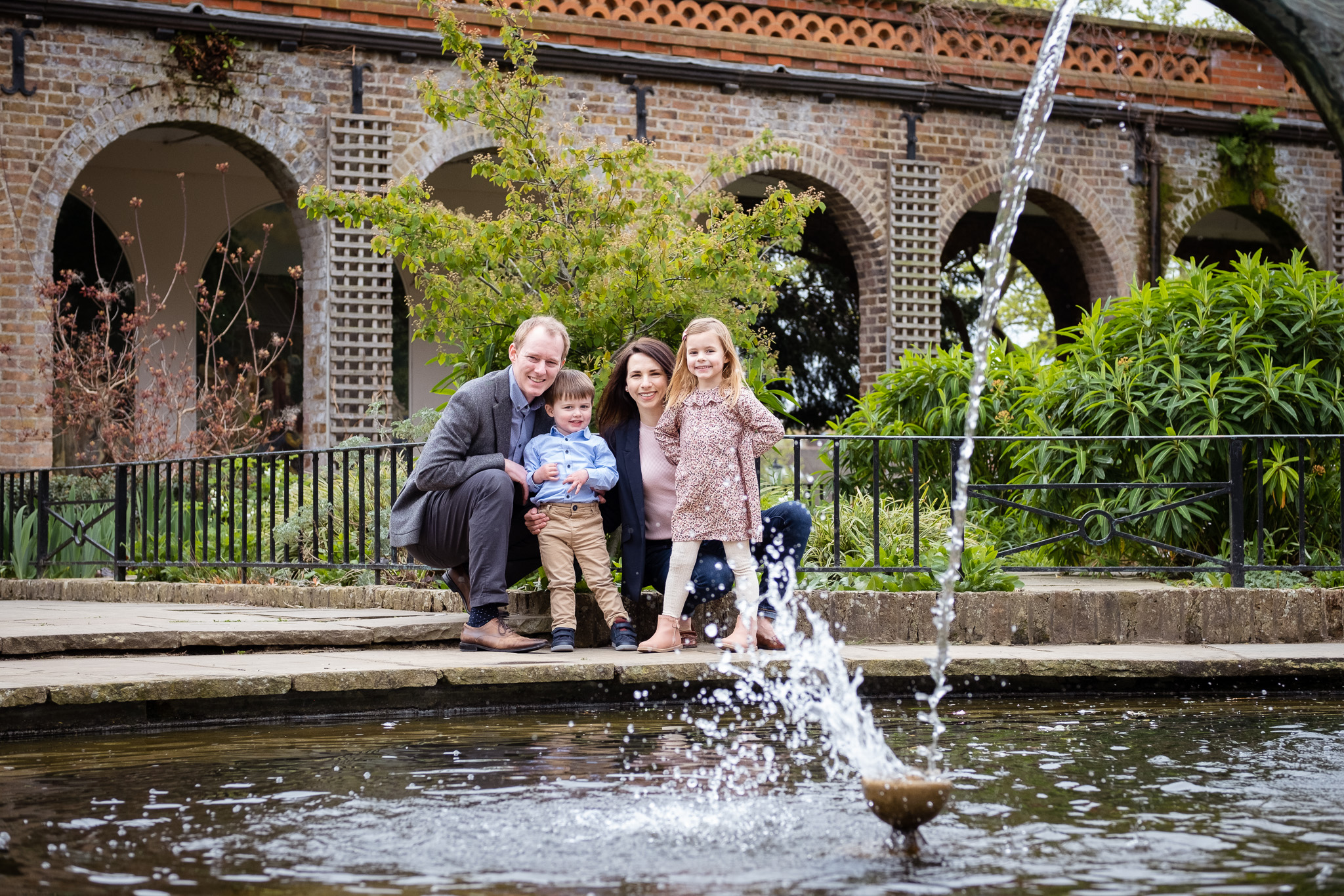 Family of four posing for a professional photo session in one of the best parks in London, there is a fountain nearby