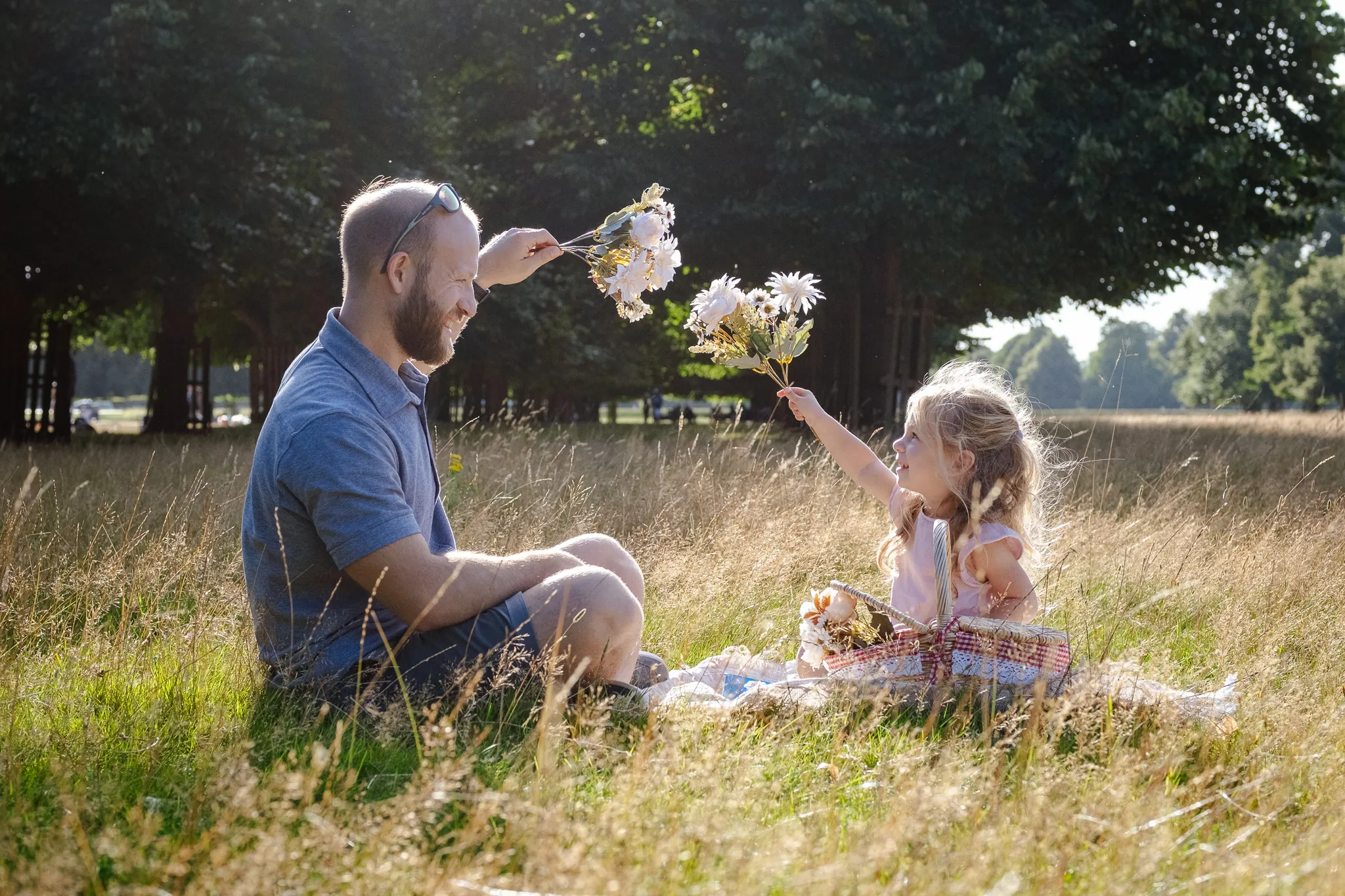 Father and daughter playing in a park, both holding flowers, sitting on a blanket, the Sun is behind them, almost sunset